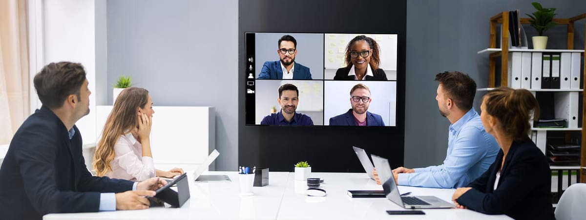 Conference room with 4 people at the table and 4 others connected via monitor and video conference