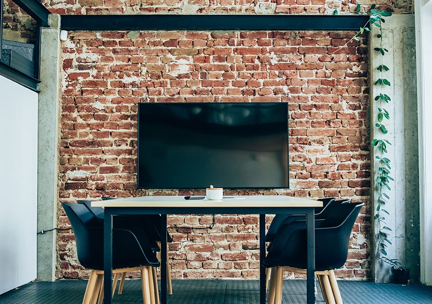 In a small meeting room, a large screen hangs on the wall in front of a table with 6 chairs