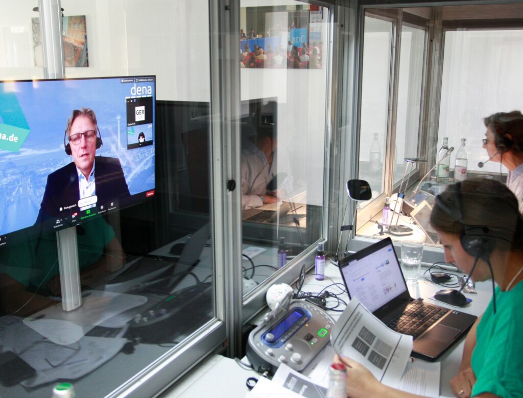Два переводчика сидят в кабине переводчика в студии синхронного перевода PCS и смотрят на большой монитор, на котором изображен говорящий во время синхронного перевода.