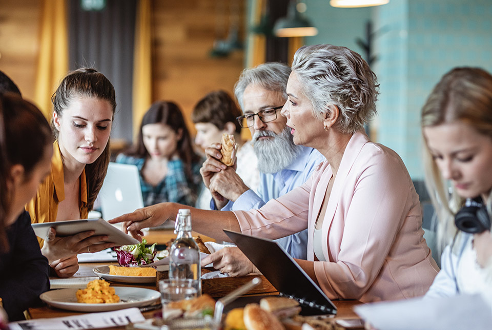 Lively meeting of about 8 people at a conference table filled with laptops, drinks, but also food, it all seems very lively and stimulating