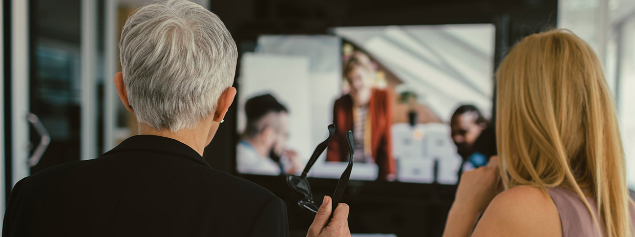 zwei Konferenzteilnehmerinnen schauen auf andere Konferenzteilnehmende, die per Monitor zugeschaltet sind