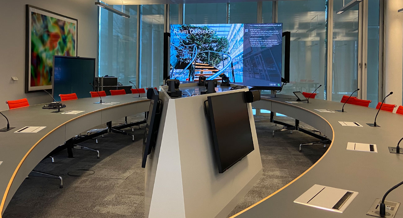 Fertige Installation eines hybriden Konferenzraumes, ein runder Tisch für 20 Personen, mit eingebauten Tischsprechstellen, in der Mitte des runden Tisches Monitore und PTZ-Kameras