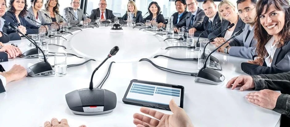 16 Personen sitzen an einem runden Tagungstisch mit Mikrofonanlage, wo jeder Teilnehmende eine eigene Sprechstelle vor sich stehen hat, um sich darüber zu Wort melden zu können. Die Anlage ist von Bosch und heißt CCS 1000 D