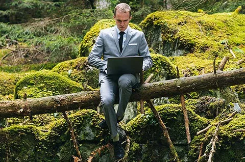 A man in business attire and tie sits on a fallen tree in a very mossy patch of forest at his laptop
