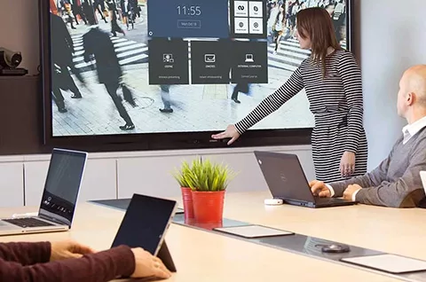 In einem kleinen Konferenzraum mit 3 Personen am Besprechungstisch steht eine Frau an einem großen Bildschirm an der Wand und stellt etwas an dem Monitor ein