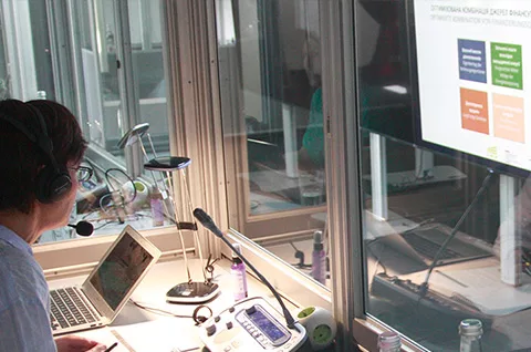 Dolmetscherin sitzt in einem Dolmetsch-Hub der Firma PCS in einer Dolmetschkabine und schaut beim Simultandolmetschen auf einen großen Monitor, der eine Präsentation zeigt.