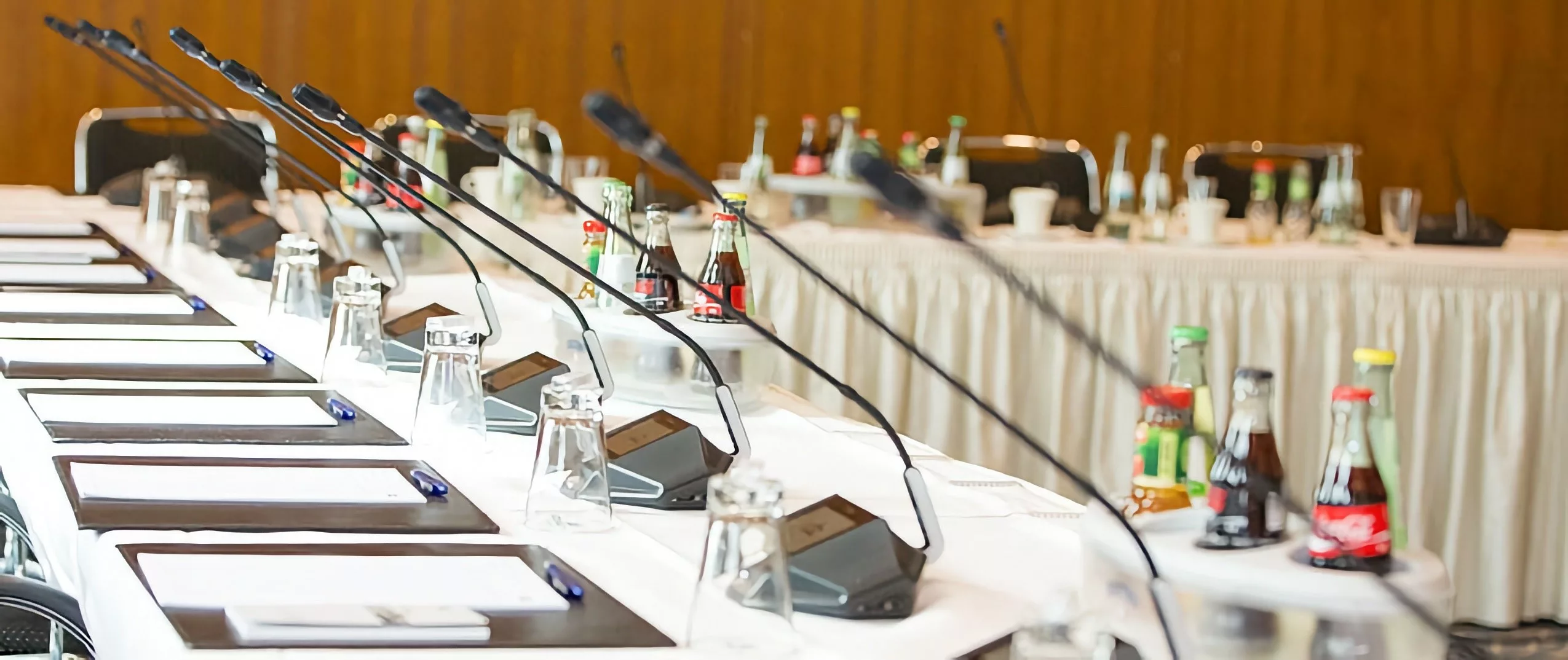 Vue sur une rangée de tables de conférence préparées pour une réunion, équipées de sous-mains, de boissons et de postes de conférence sans fil - tout semble prêt pour une conférence.